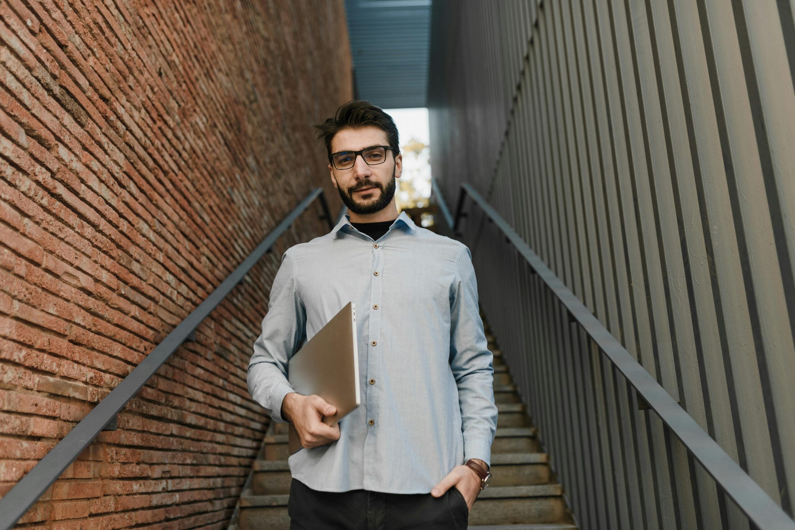 Man in eyeglasses holding laptop on stairs between brick and metal walls.